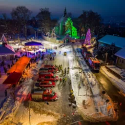 Nocna panorama zimowa: Rynek miejski z oświetlonym kościołem, choinką i sceną, straż pożarna w akcji, widziana z góry.