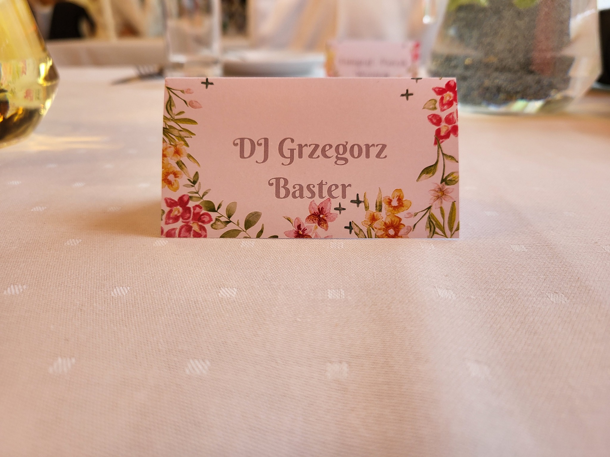 Winietka z imieniem DJ Grzegorz Baster, ozdobiona kwiatowym motywem, ustawiona na stole z kremowym obrusem w kropki, w tle naczynia i sztućce.