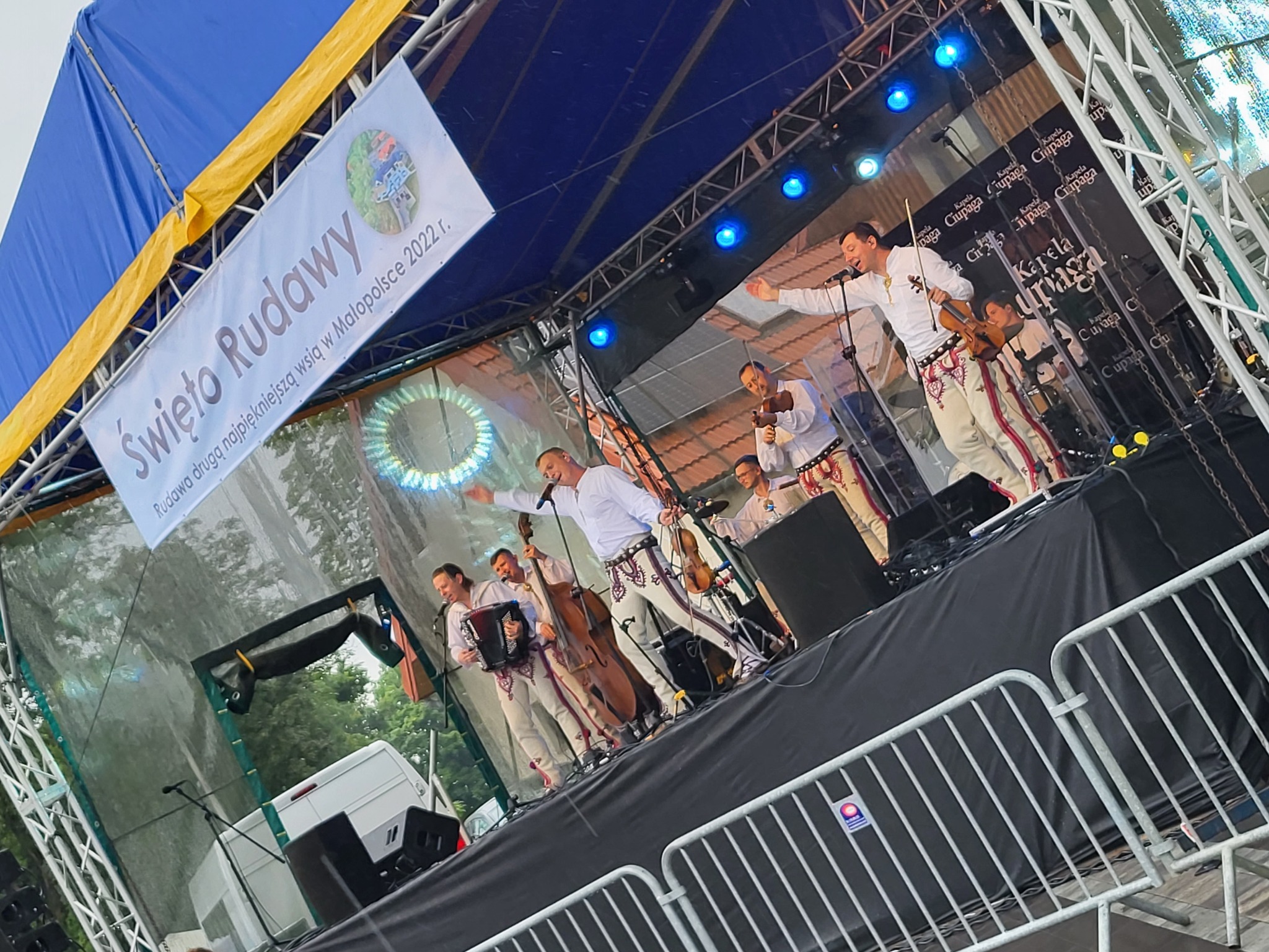 Występ kapeli ludowej na scenie plenerowej podczas festynu 'Święto Rudawy', muzycy w strojach regionalnych grają na skrzypcach, akordeonie i kontrabasie, oświetlenie sceniczne, baner reklamowy w tle.