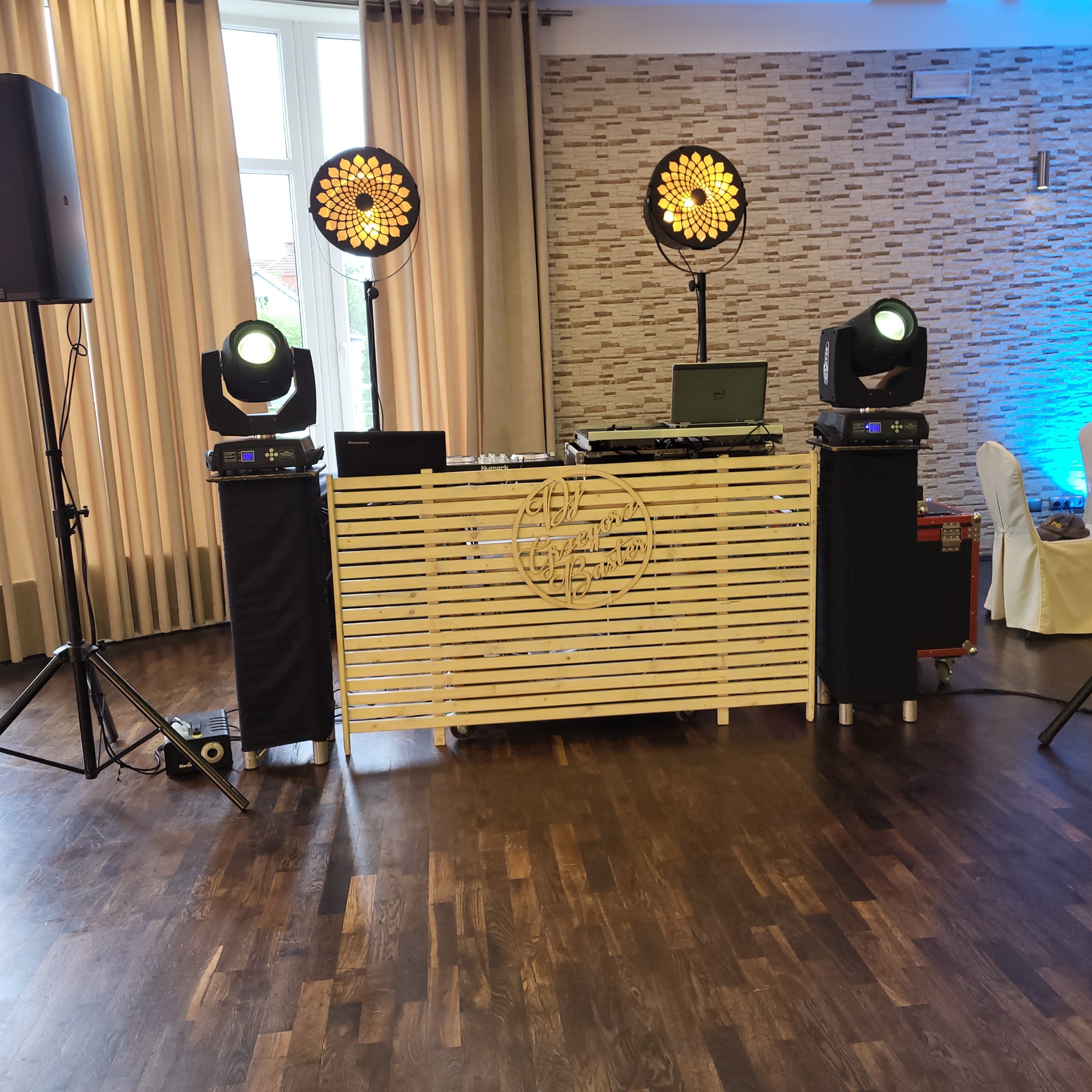 Sprzęt nagłośnieniowy i oświetleniowy przygotowany do występu w sali bankietowej, widoczne reflektory, kolumny głośnikowe, mikser i drewniana zabudowa z napisem 'DJ Gregory Buster'.
