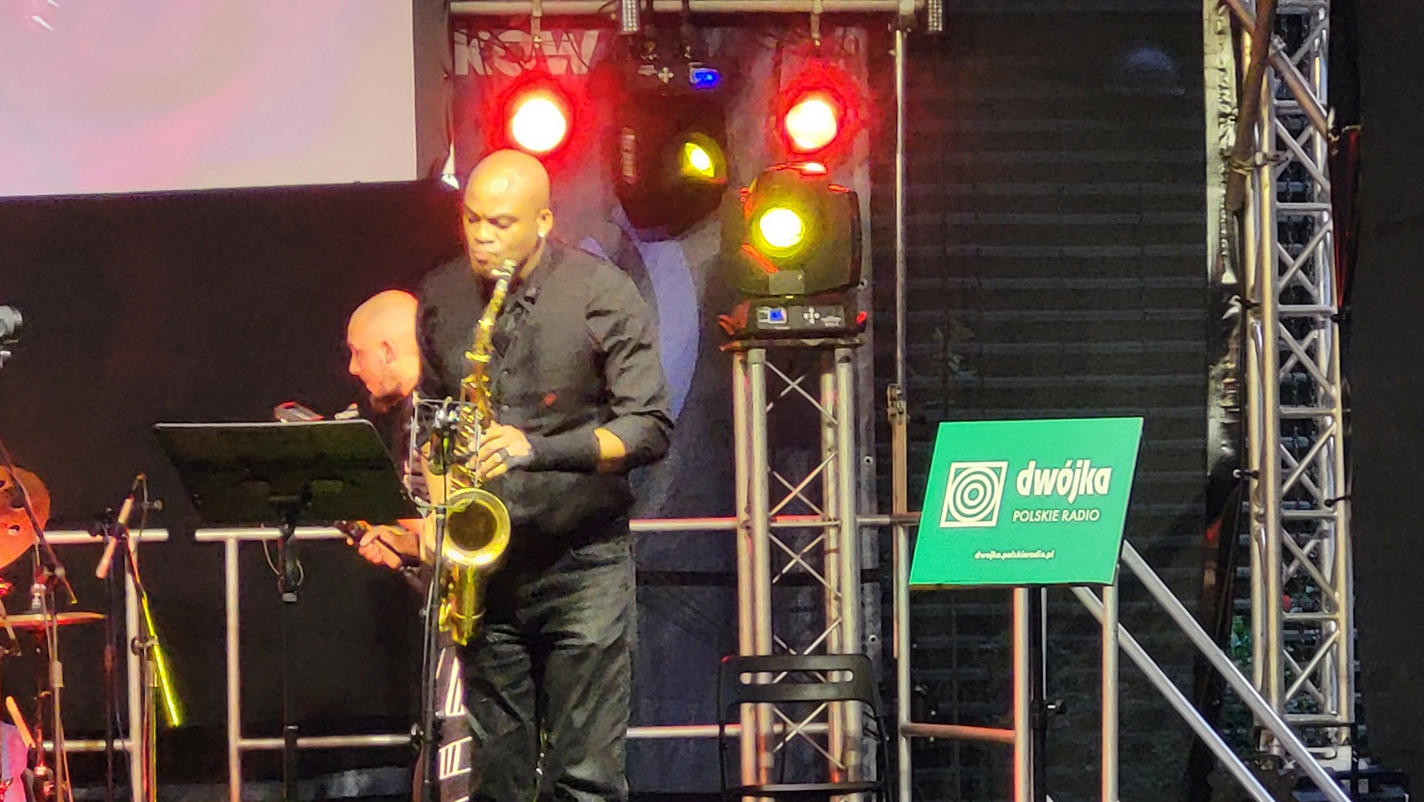 Saksofonista w czarnej koszuli gra na saksofonie podczas koncertu na scenie z oświetleniem i logo Dwójki Polskie Radio w tle.