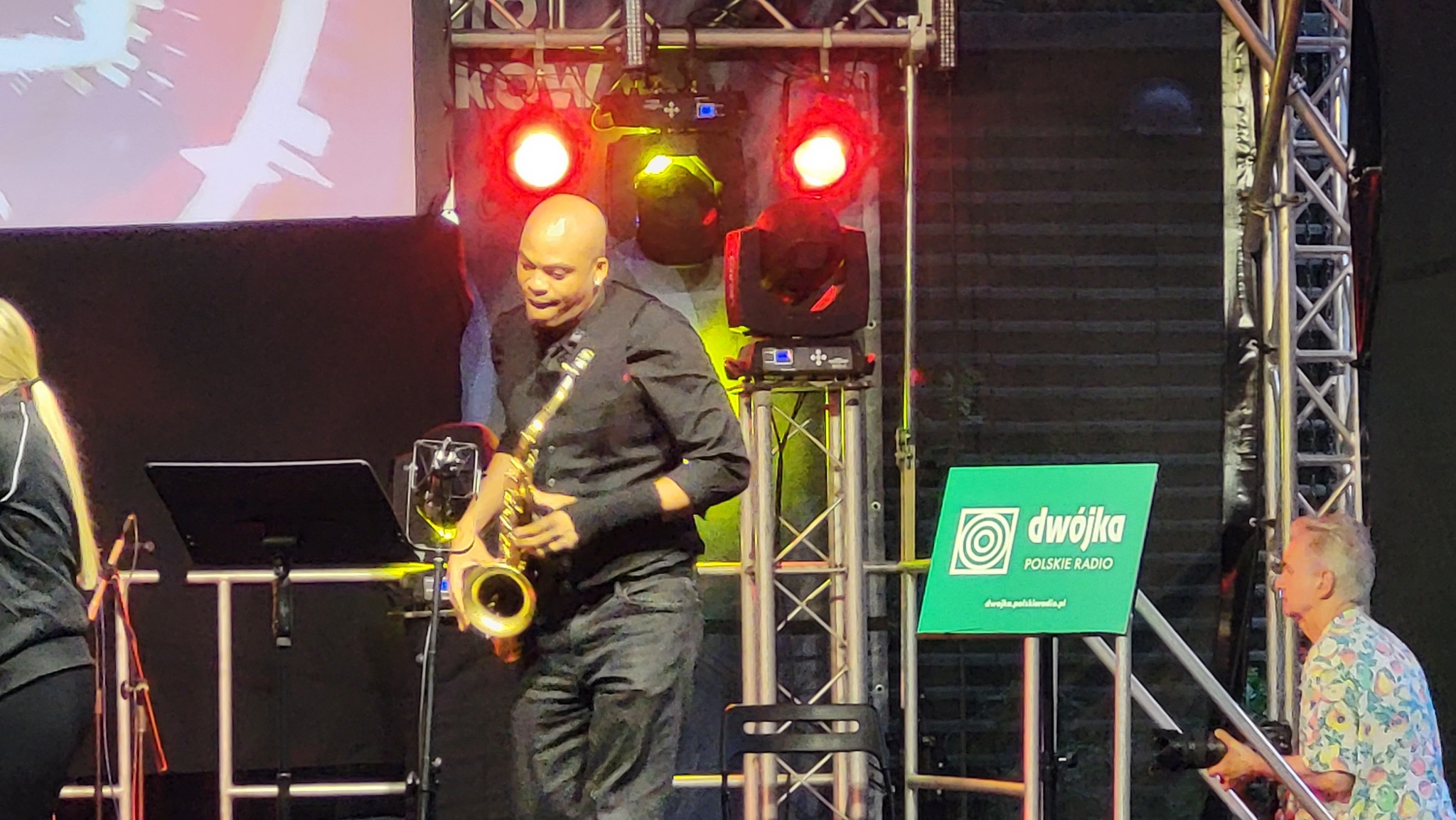 Saksofonista w czarnej koszuli gra na saksofonie na scenie oświetlonej czerwonymi reflektorami, obok widoczny znak Polskiego Radia Dwójka i konstrukcja sceniczna.