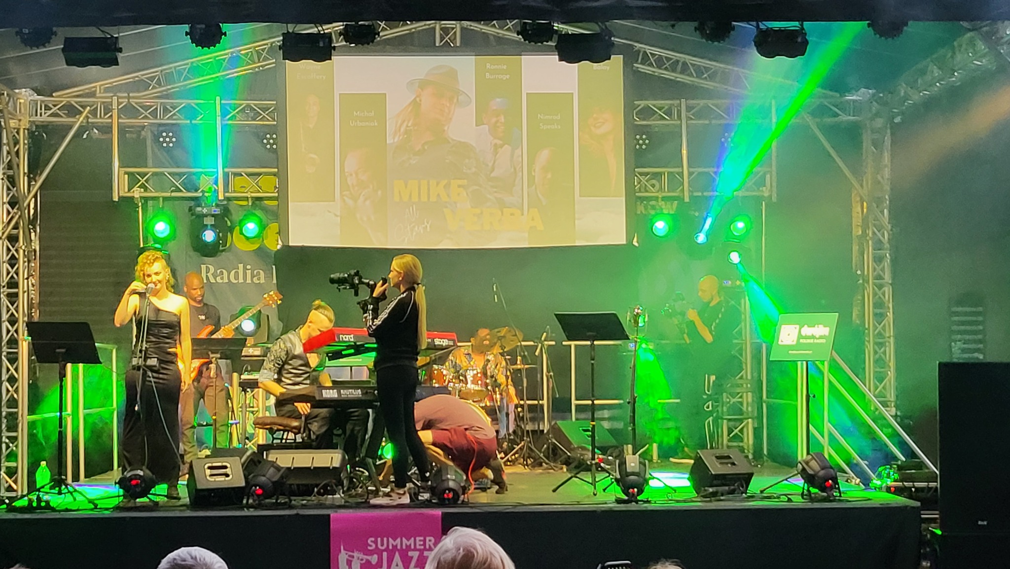 Scena plenerowa podczas festiwalu jazzowego, wokalistka w czarnej sukni śpiewa do mikrofonu, w tle muzycy grają na instrumentach, oświetlenie sceniczne w kolorze zielonym, na ekranie wyświetlają...