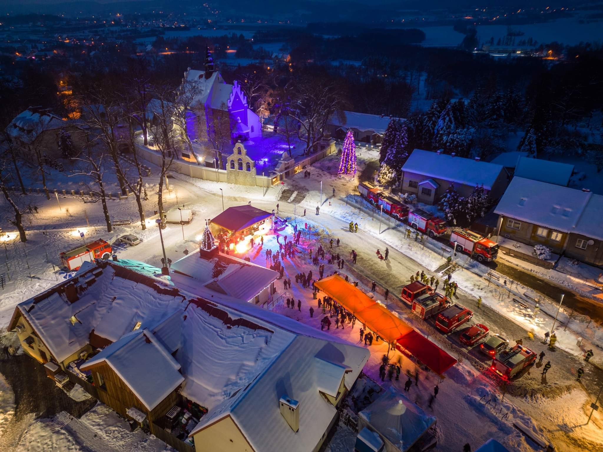 Widok z lotu ptaka na zimowy wieczór z oświetloną sceną, kościołem w fioletowym świetle i wozami strażackimi w tle.