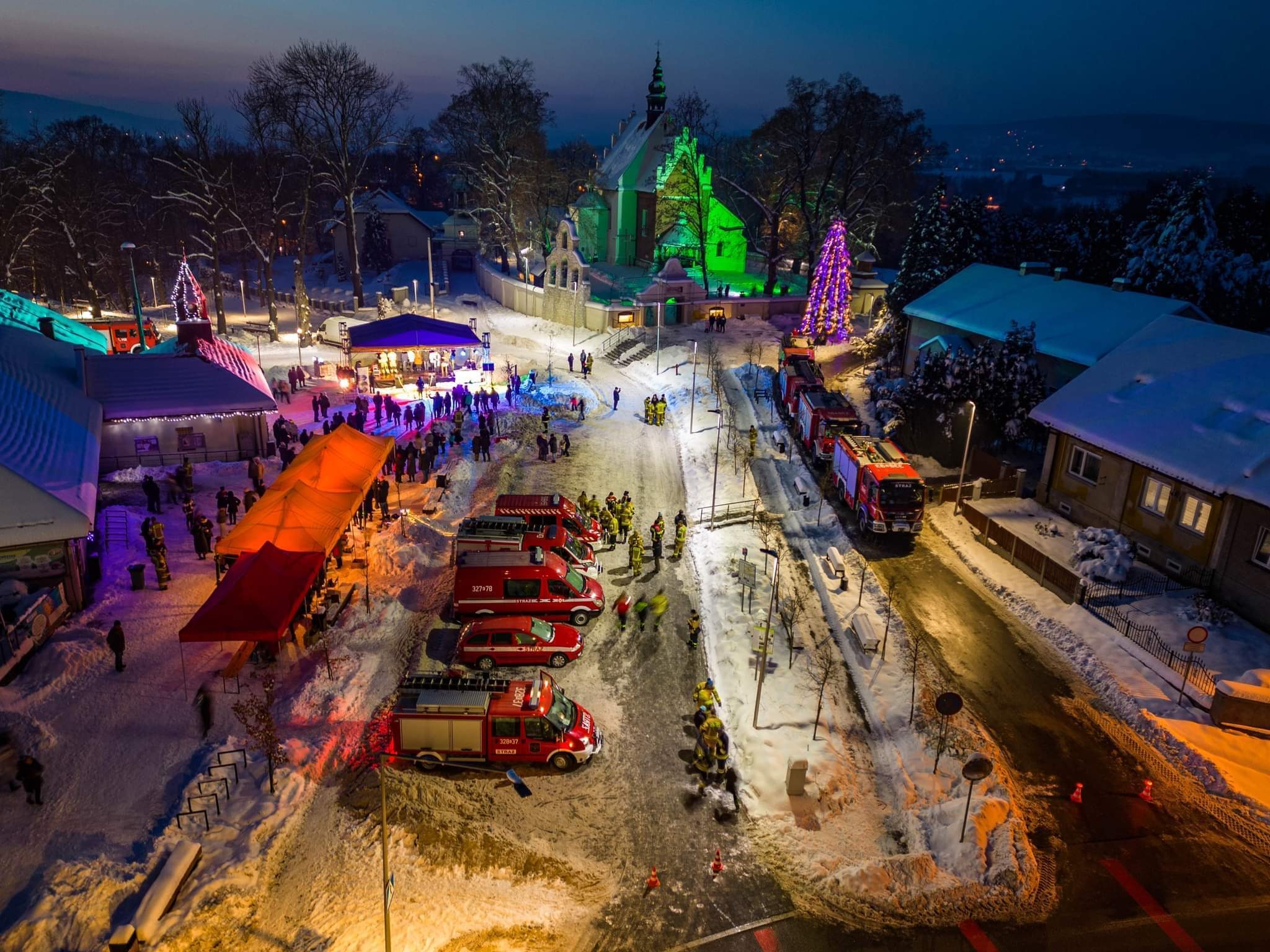 Nocna panorama zimowa: Rynek miejski z oświetlonym kościołem, choinką i sceną, straż pożarna w akcji, widziana z góry.