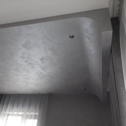 Usługi budowlane - Szara, strukturalna powierzchnia sufitu z zaokrąglonym narożnikiem i dwoma wbudowanymi punktami świetlnymi, widok z dołu, obok okno z firankami.