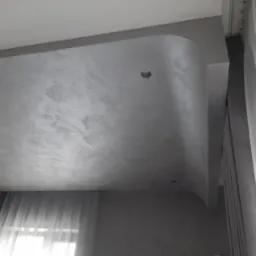 Szara, strukturalna powierzchnia sufitu z zaokrąglonym narożnikiem i dwoma wbudowanymi punktami świetlnymi, widok z dołu, obok okno z firankami.