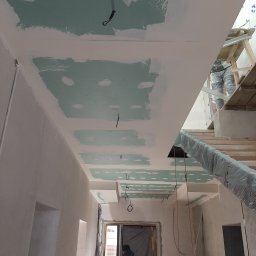 Usługi budowlane - Wnętrze w trakcie prac wykończeniowych: sufit z płyt gipsowo-kartonowych częściowo pokryty gładzią, widoczne przewody elektryczne, fragment schodów i pracownicy w tle.