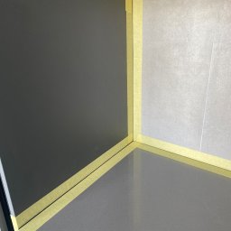 Usługi budowlane - Przygotowanie do malowania: fragment ściany i blatu zabezpieczone żółtą taśmą malarską, widoczne dwie różne powierzchnie - ciemnoszara ściana i jasnoszara płytka.