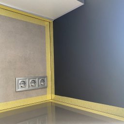 Usługi budowlane - Przygotowanie do malowania: ściana pomalowana na ciemny kolor, obok ściana z szarymi płytkami, zabezpieczone taśmą malarską, widoczne gniazdka elektryczne.