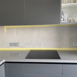 Usługi budowlane - Przygotowanie do malowania ściany nad blatem kuchennym: zabezpieczone taśmą malarską płytki i blat, widoczne gniazdka elektryczne.