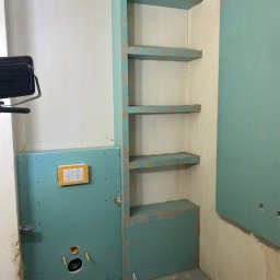 Usługi budowlane - Niedokończona łazienka z płytami karton-gips: zabudowa stelaża WC i wnęka z półkami z płyty GK, widoczne instalacje wodno-kanalizacyjne, przygotowanie do dalszych prac wykończeniowych.