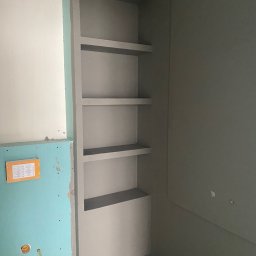 Usługi budowlane - Wnęka z półkami w surowym stanie w łazience, ściany przygotowane pod malowanie, widoczne elementy instalacji hydraulicznej na ścianie z płyt gipsowych.