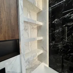 Wykończona łazienka z białymi i czarnymi marmurowymi płytkami, wnęką z półkami podświetloną LED, oraz fragmentem drewnianej szafki.