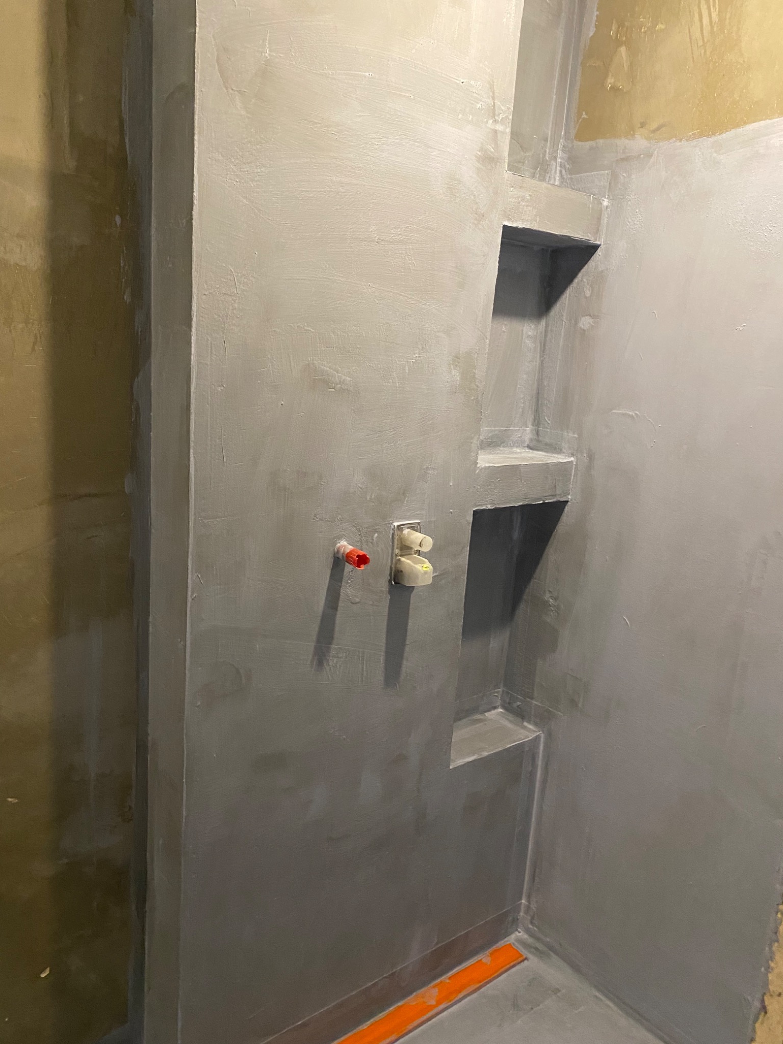 Przygotowana do dalszych prac łazienka z szarymi ścianami, wnękami półkowymi i widocznymi instalacjami wodnymi z wystającymi elementami z tworzywa w kolorze czerwonym i białym.