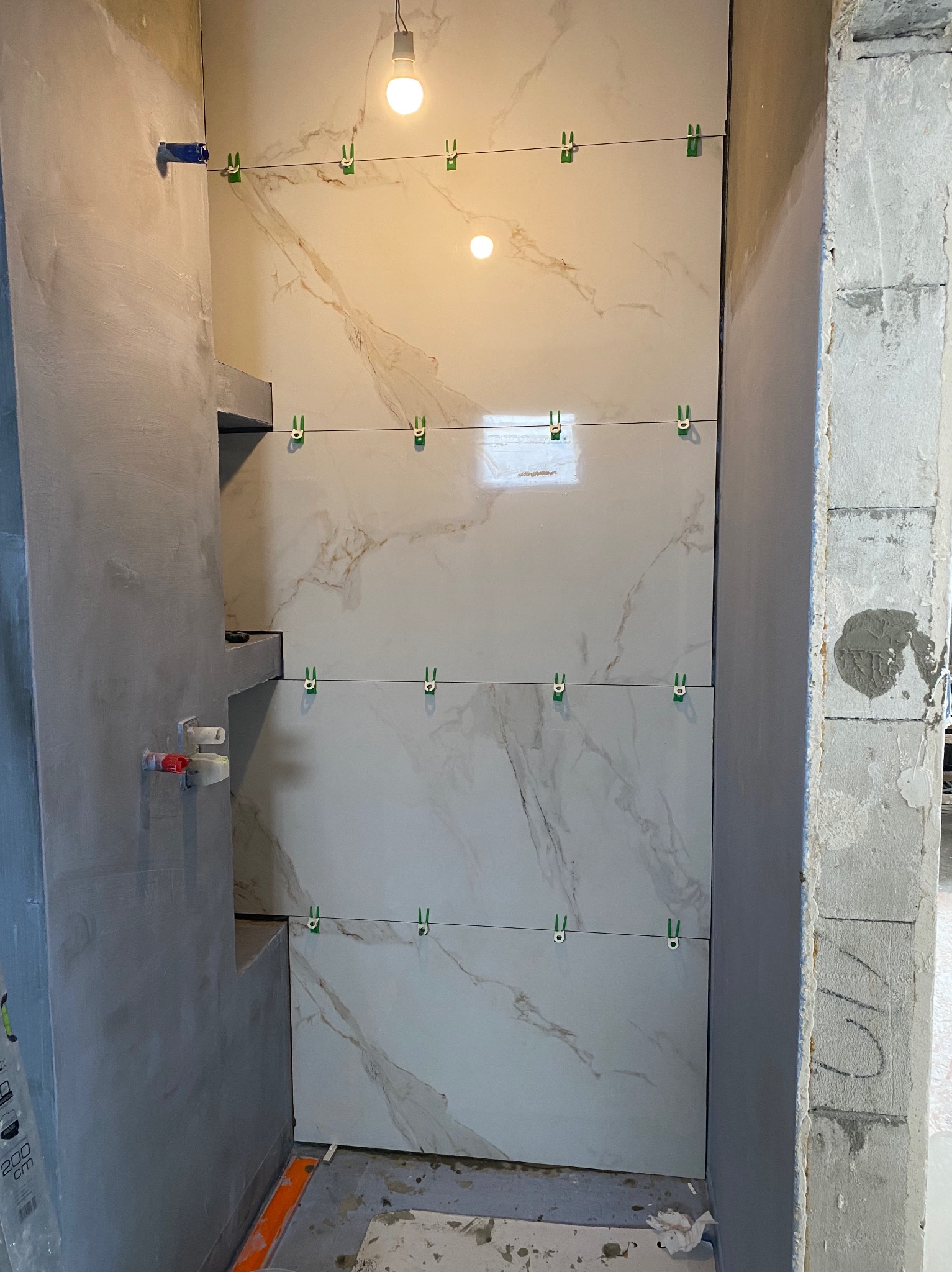Montaż płytek imitujących marmur w łazience, widoczne zielone klipsy poziomujące, surowa ściana z betonu komórkowego i oświetlenie żarówkowe.