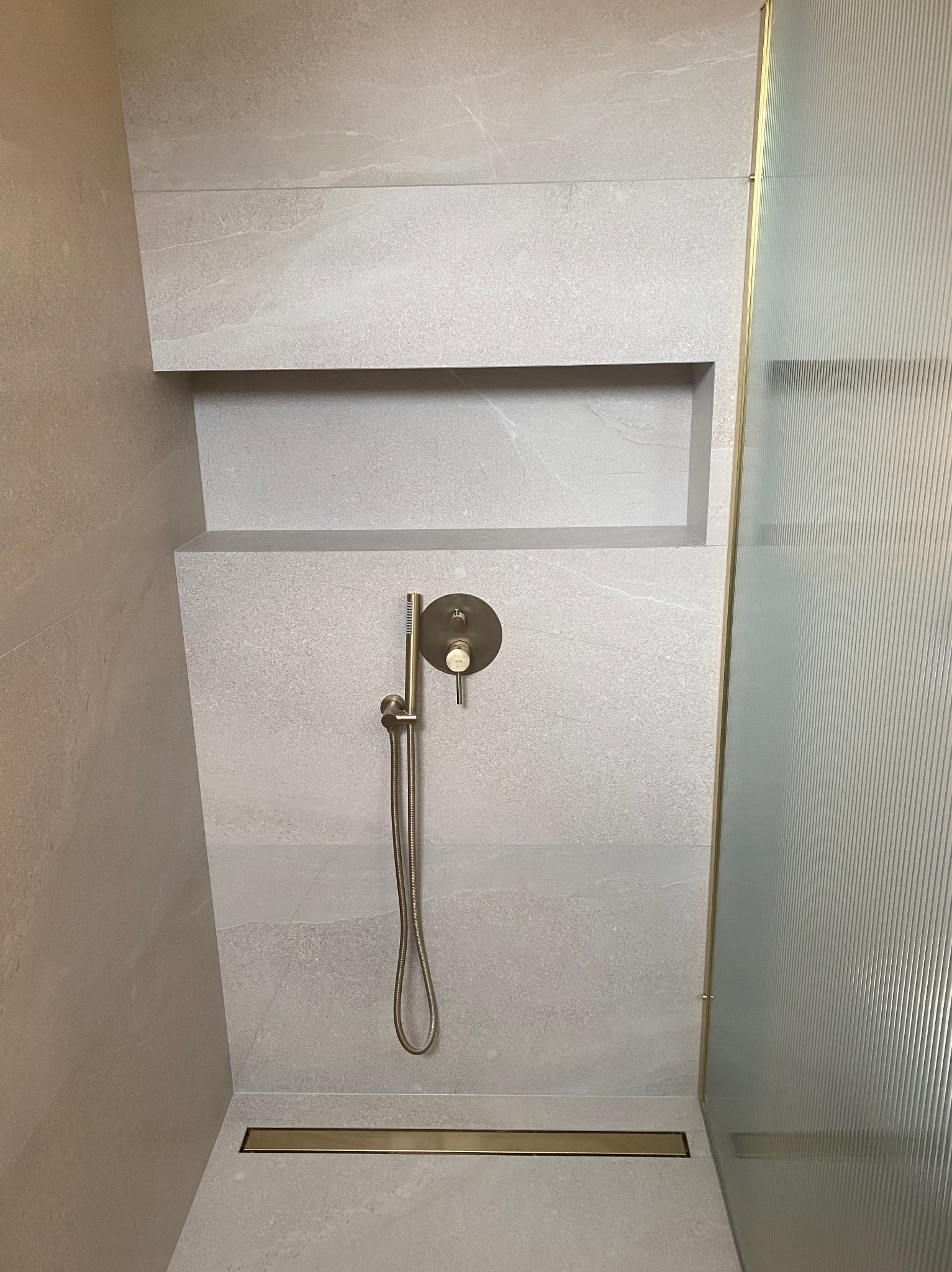 Wykończona łazienka: szara płytka imitująca kamień, wnęka w ścianie, złota armatura prysznicowa i odpływ liniowy, ryflowana szyba kabiny prysznicowej.