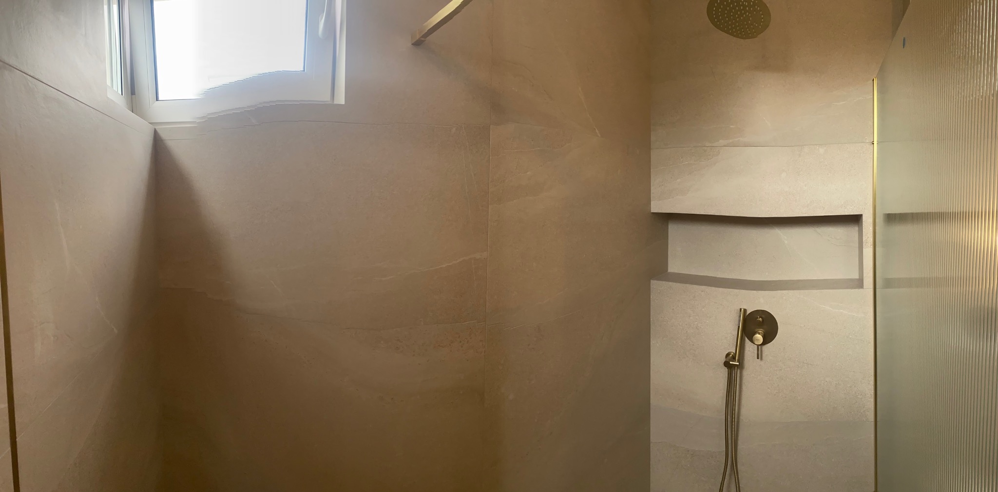 Beżowa łazienka z kabiną prysznicową typu walk-in, wnęką w ścianie i armaturą w kolorze złotym.