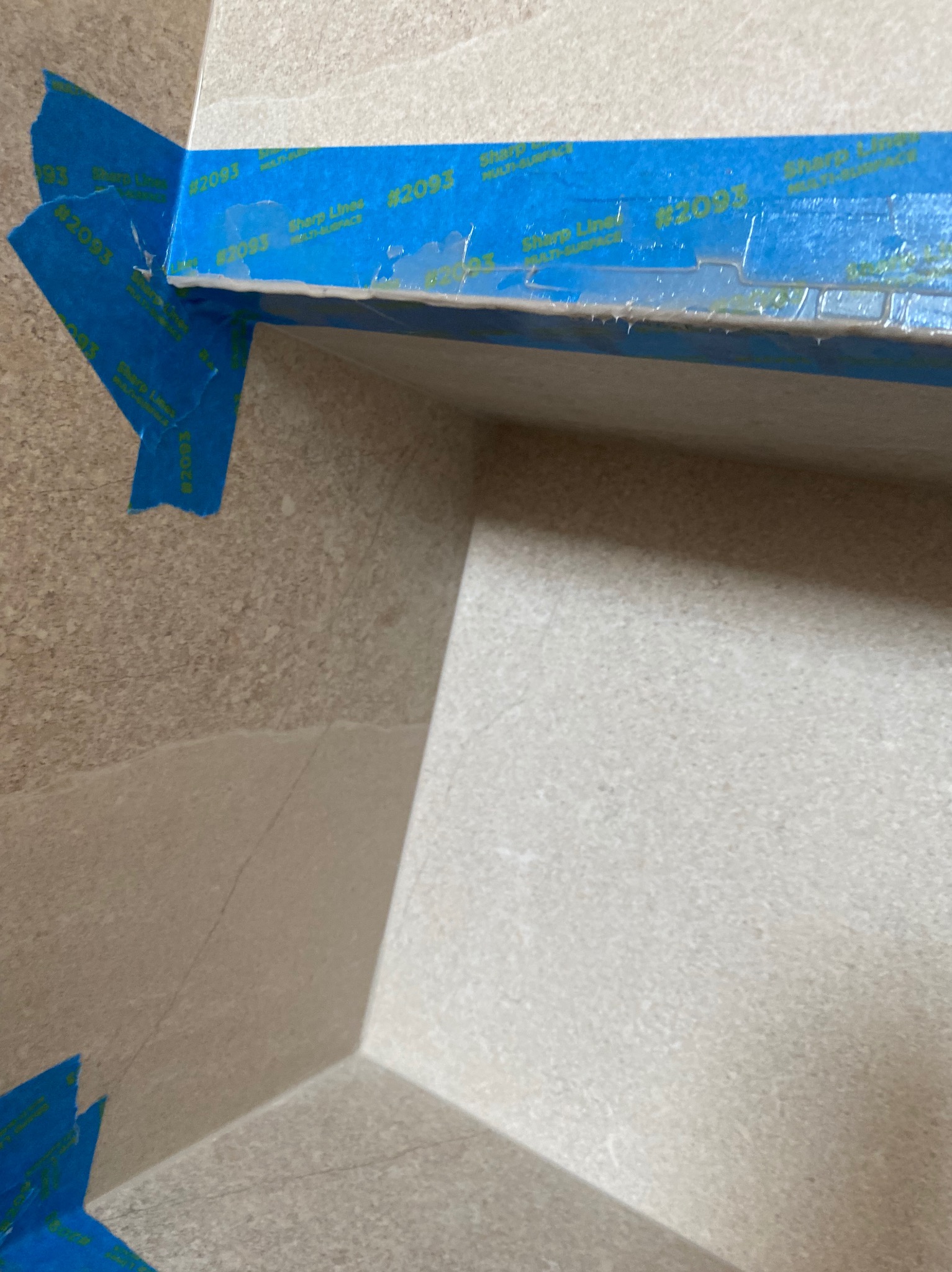 Fragment narożnika obłożonego płytkami, zabezpieczonego niebieską taśmą malarską Sharp Lines #2093, widoczne niedoskonałości i pęknięcia na powierzchni płytek.