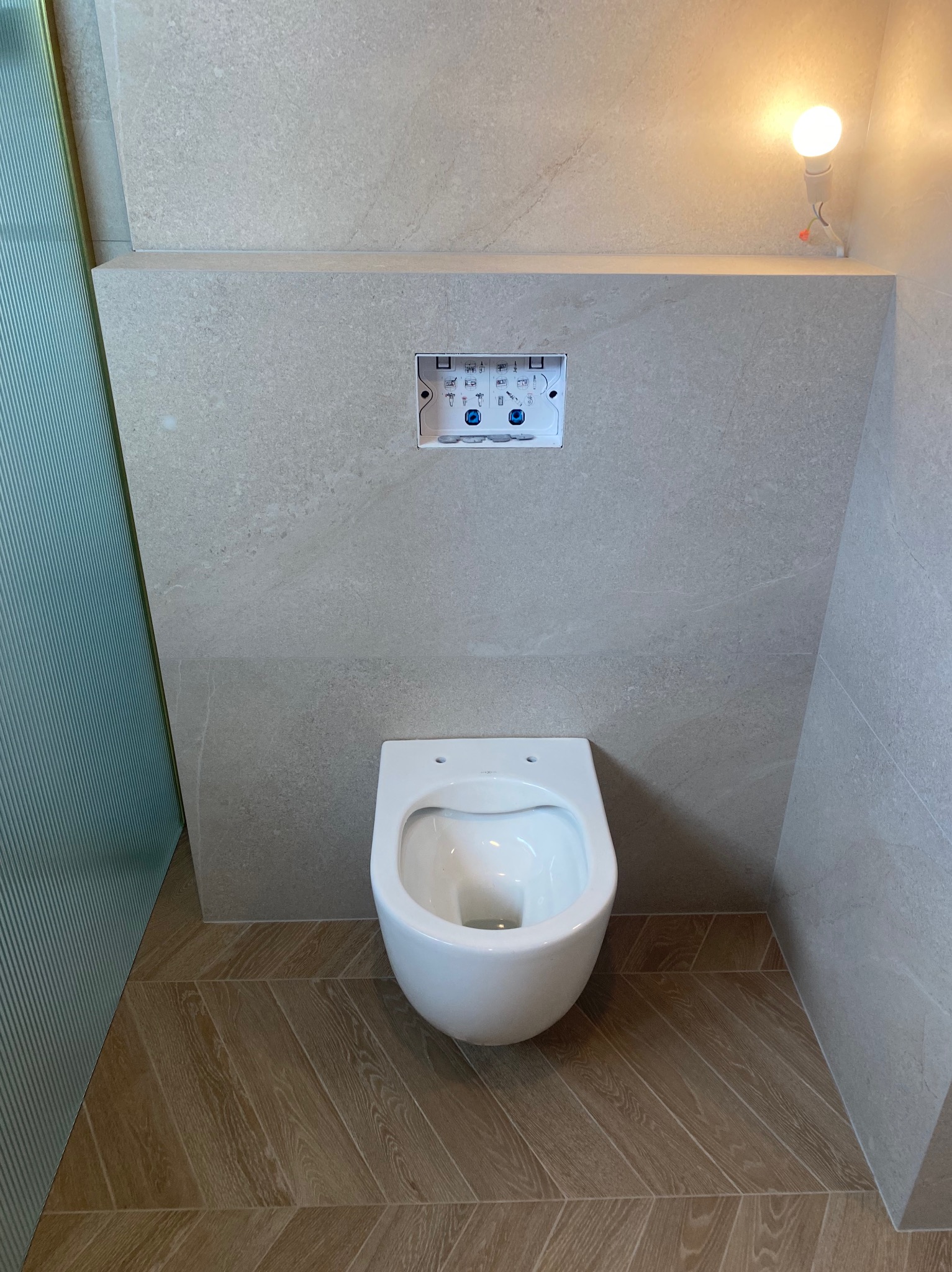 Podwieszana miska WC z zabudowanym stelażem i przyciskiem spłukującym, wykończona płytkami w odcieniach beżu i brązu z drewnopodobną podłogą ułożoną w jodełkę.