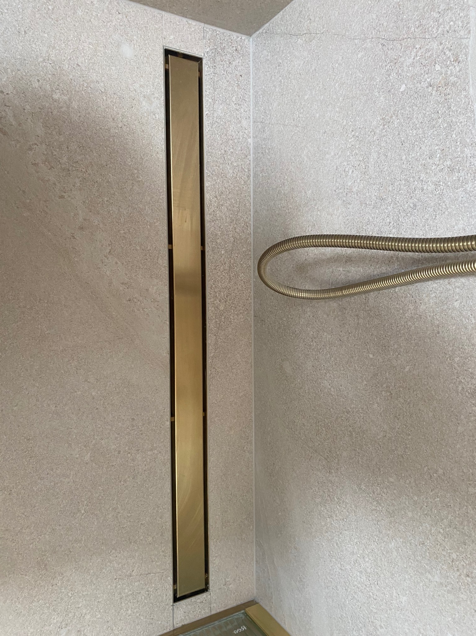 Beżowa ściana łazienki z pionowym, złotym odpływem liniowym i zwisającym, złotym wężem prysznicowym.