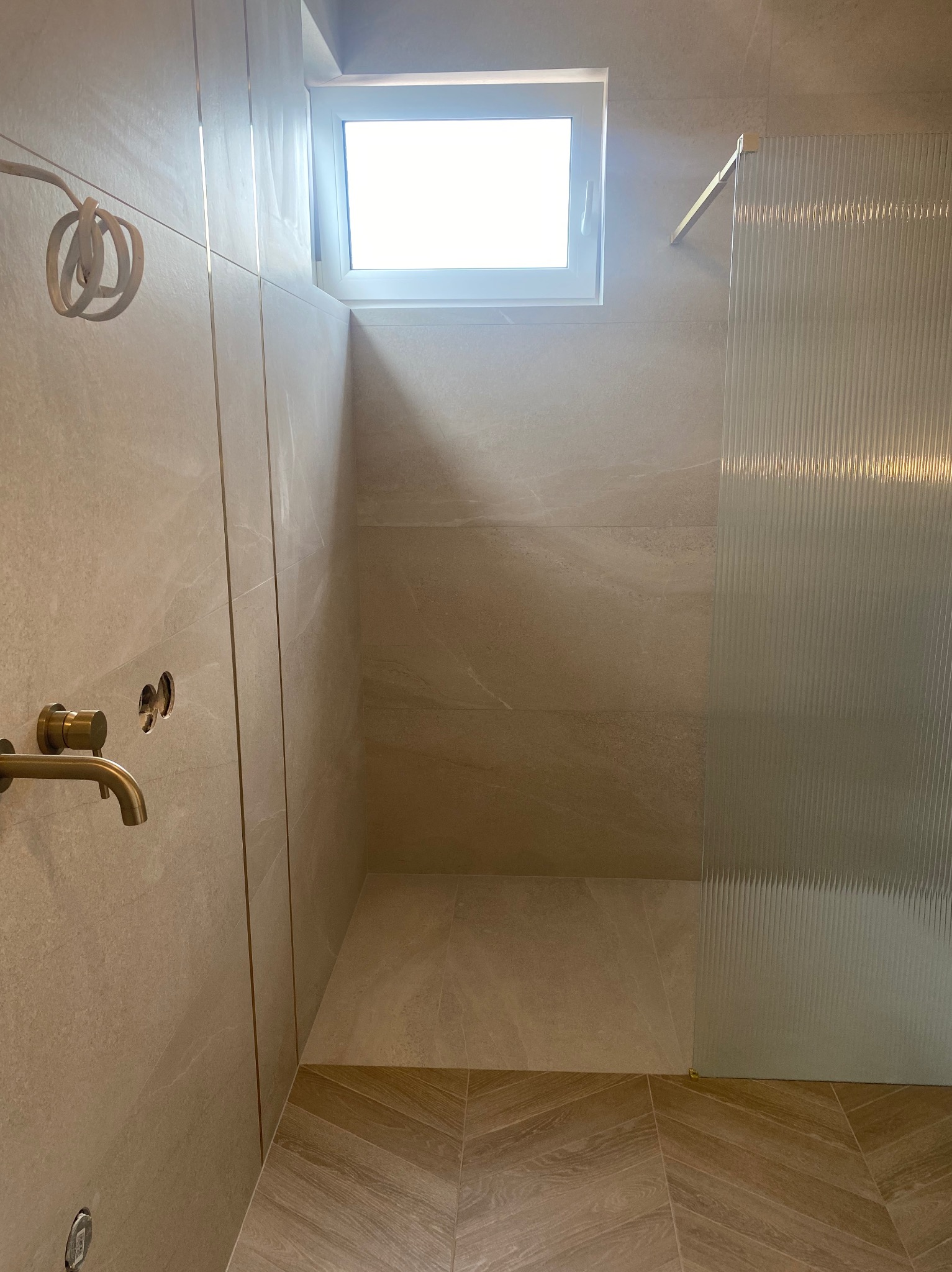 Nowoczesna łazienka w beżowych odcieniach z prążkowaną szybą prysznicową, złotą baterią i niedokończonym okablowaniem.