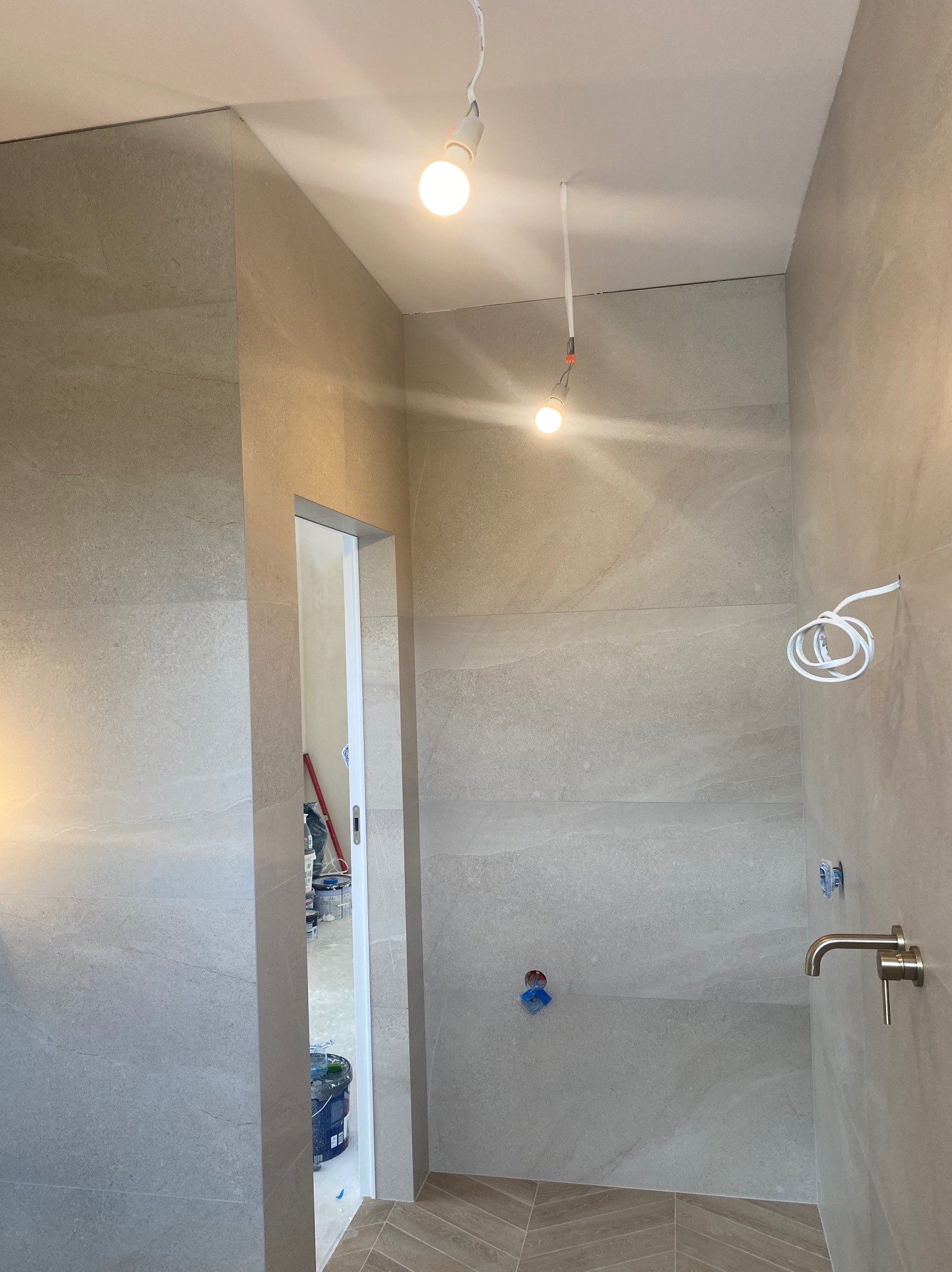 Łazienka w trakcie wykańczania: położone płytki imitujące kamień na ścianach i podłodze w jodełkę, widoczne wyprowadzenia instalacji wodnej i elektrycznej, surowe żarówki zwisające z sufitu, drzwi...