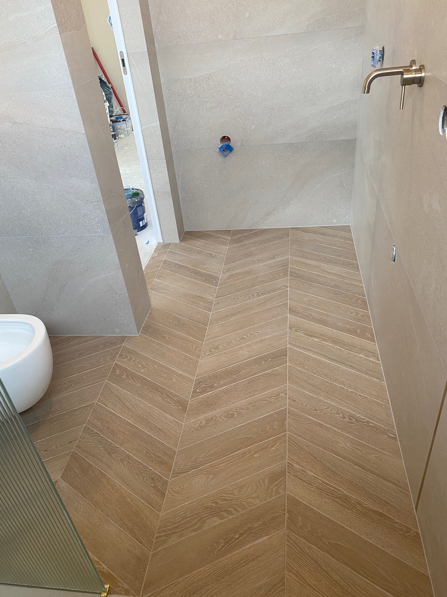 Nowoczesna łazienka w trakcie wykańczania: beżowe płytki na ścianach i podłodze ułożone we wzór jodełki, widoczna armatura, fragment toalety i kabiny prysznicowej. W tle widoczne pojemniki z farbą.