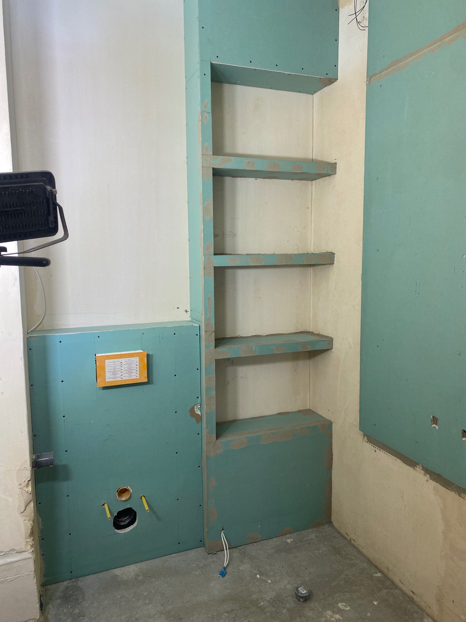 Niedokończona łazienka z płytami karton-gips: zabudowa stelaża WC i wnęka z półkami z płyty GK, widoczne instalacje wodno-kanalizacyjne, przygotowanie do dalszych prac wykończeniowych.