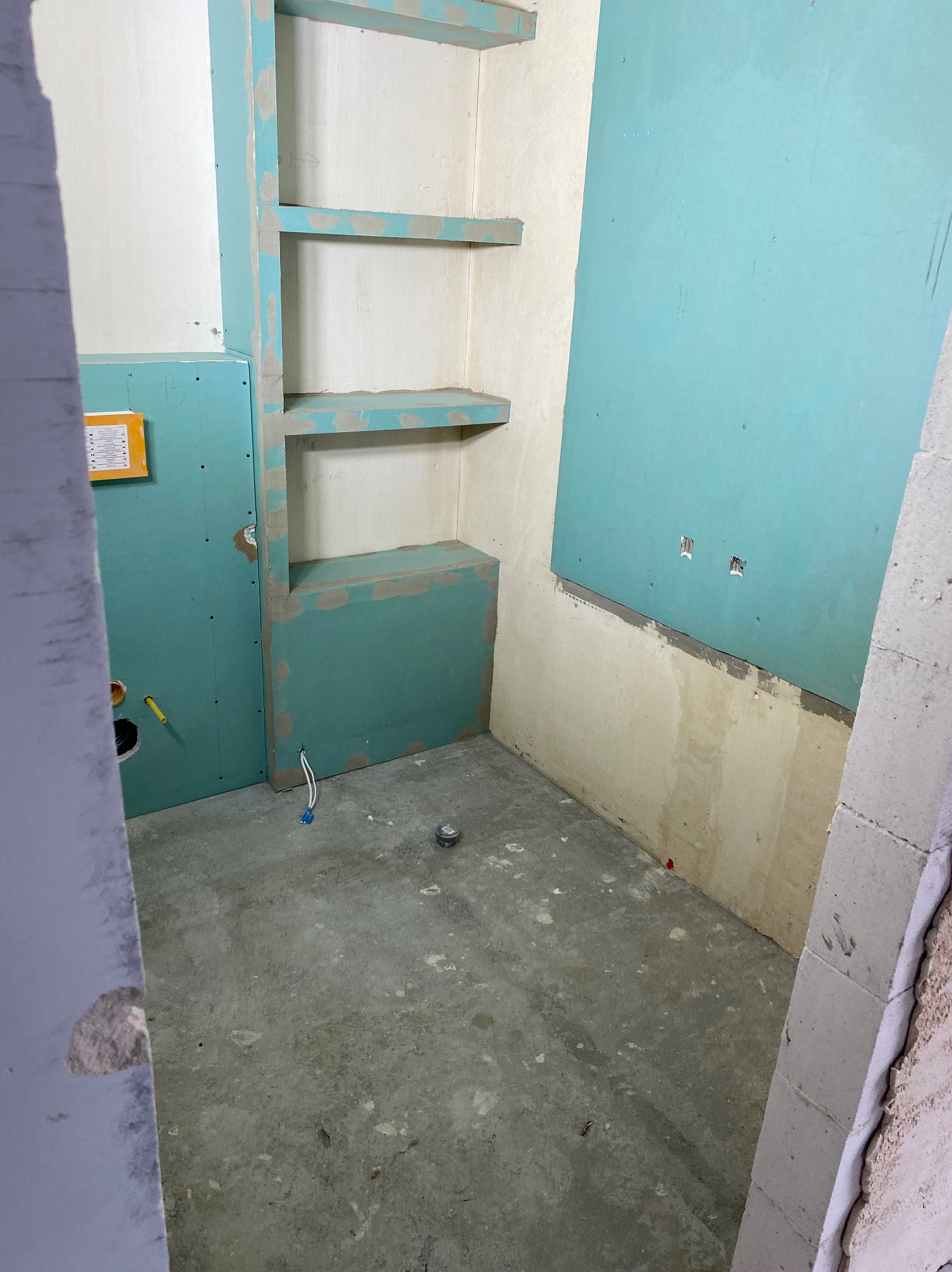 Surowy stan łazienki w trakcie prac: płyty kartonowo-gipsowe w kolorze turkusowym na ścianach, wnęka z półkami w zabudowie, widoczne instalacje elektryczne i hydrauliczne, surowy beton na podłodze.