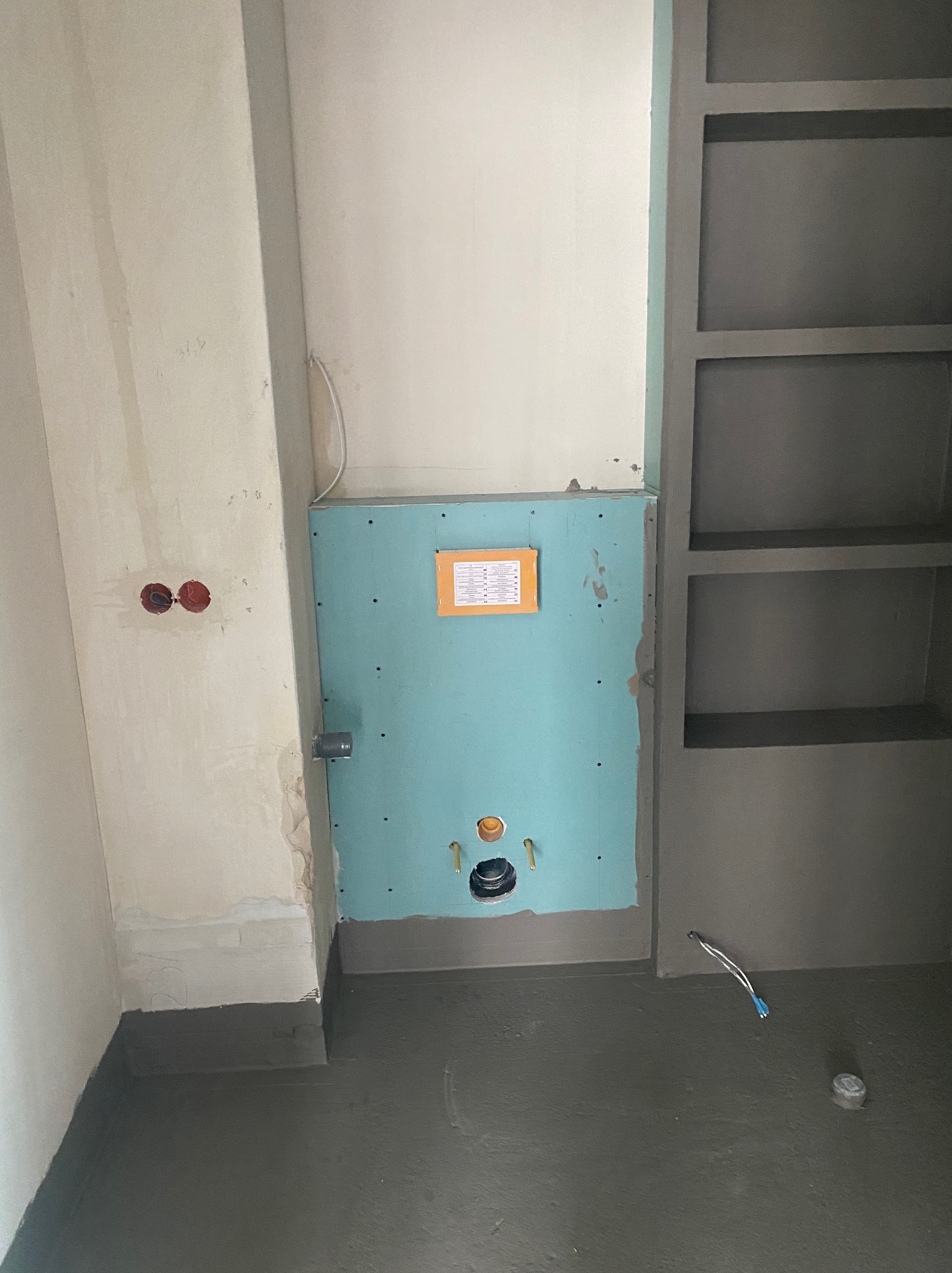 Przygotowana ściana z płyt karton-gips w łazience, widoczne otwory na instalację hydrauliczną, świeżo wylany szary podkład podłogowy, obok regał wnękowy z półkami, ściany w stanie surowym.