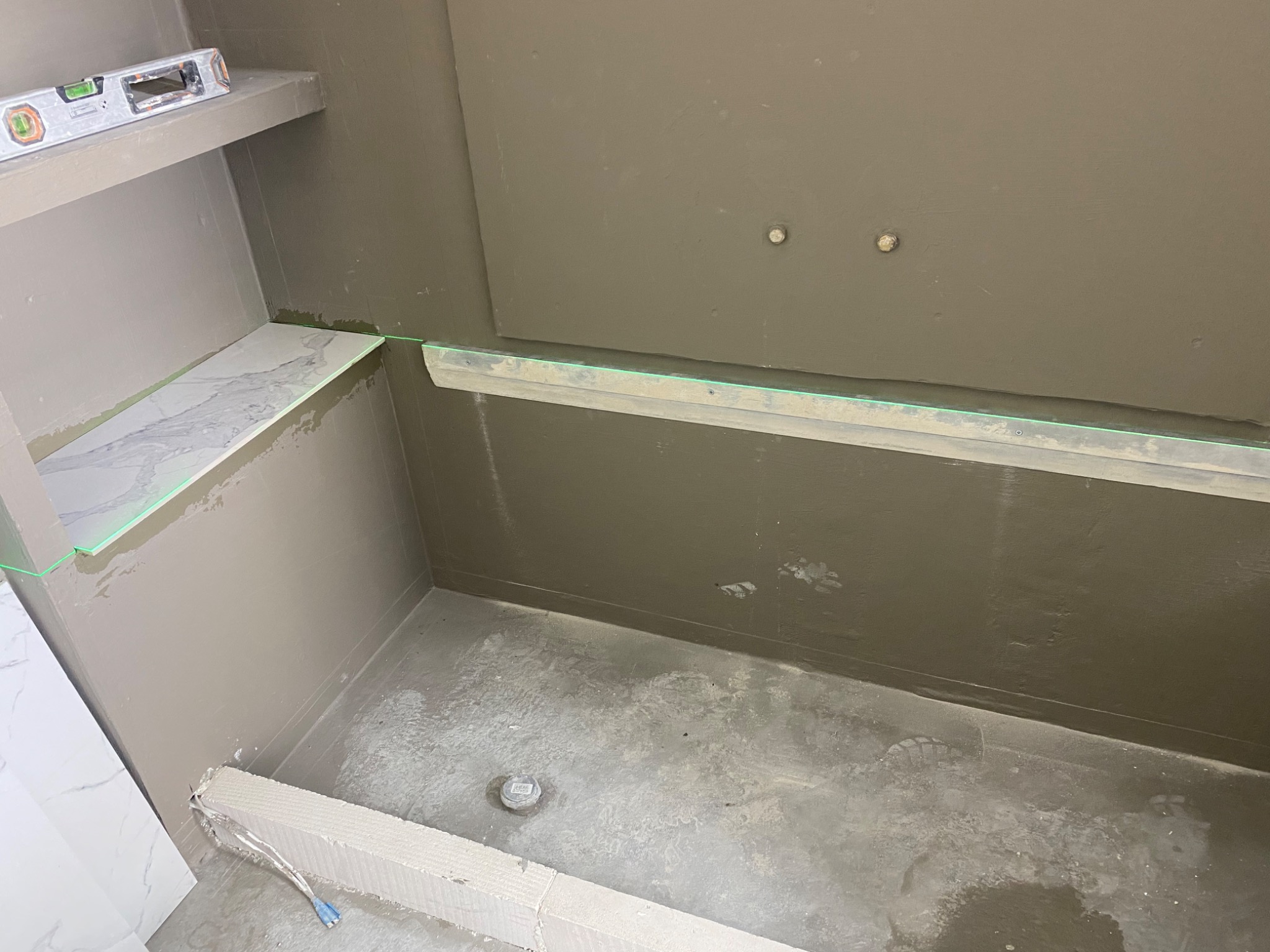 Przygotowanie do położenia płytek w łazience: hydroizolacja ścian i podłogi, poziomica laserowa wyznacza linię, półka z marmurowym blatem, widoczne ślady robocze.