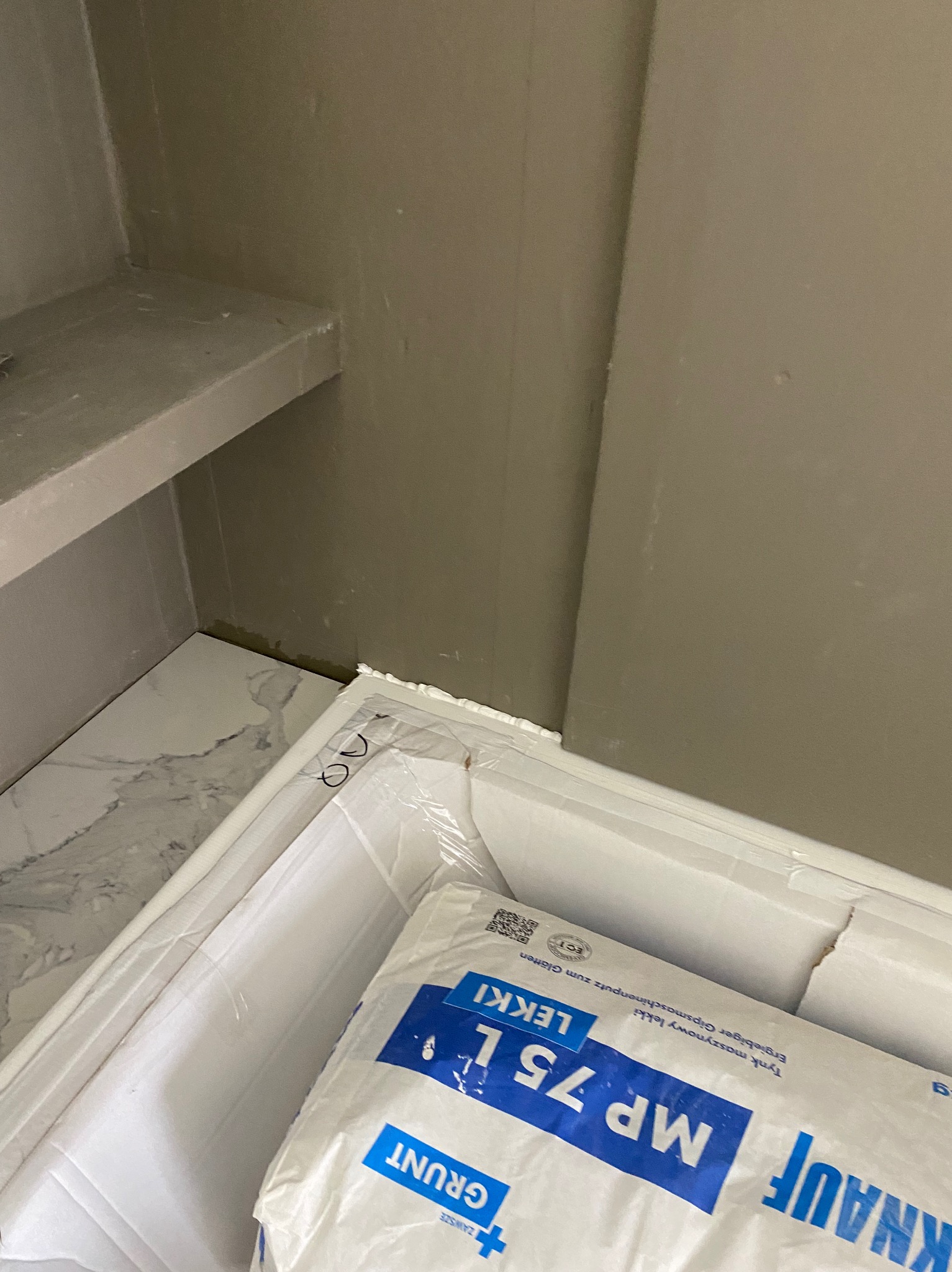 Wnętrze łazienki w trakcie prac, widoczna półka, marmurowe płytki podłogowe i worek z tynkiem maszynowym Knauf MP 75L, co wskazuje na etap przygotowania ścian do dalszych prac wykończeniowych.