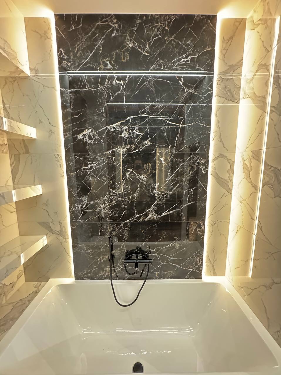 Wanna z czarną armaturą na tle ściany wyłożonej marmurowymi płytkami w odcieniach bieli i szarości, z podświetleniem LED po bokach i w zagłębieniu w ścianie.