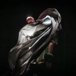 Iluzjonista w trakcie występu, rzuca płachtą materiału, w tle widoczne krzesło z zieloną tapicerką.