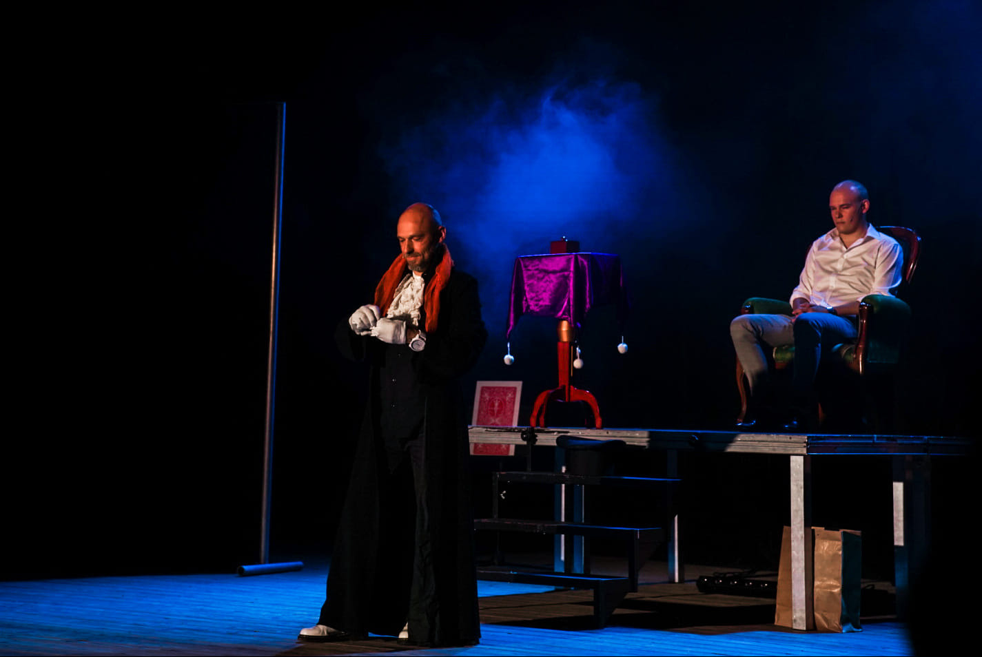 Iluzjonista w czarnym płaszczu i białych rękawiczkach przygotowuje się do występu na scenie z niebieskim oświetleniem, obok stół z rekwizytami zakryty fioletową tkaniną i siedzący widz w fotelu.