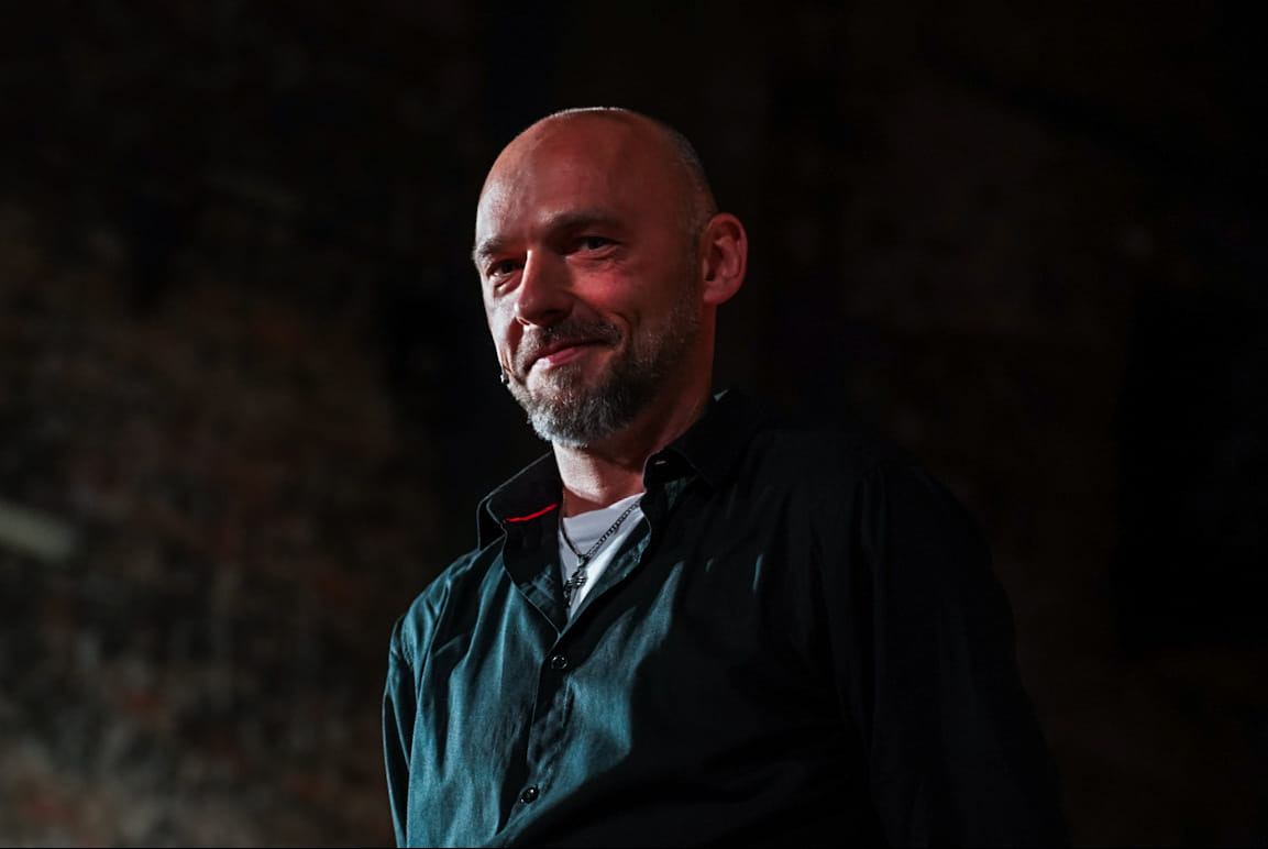 Portret uśmiechniętego iluzjonisty z mikroportem, w czarnej koszuli, na ciemnym tle ceglanej ściany.