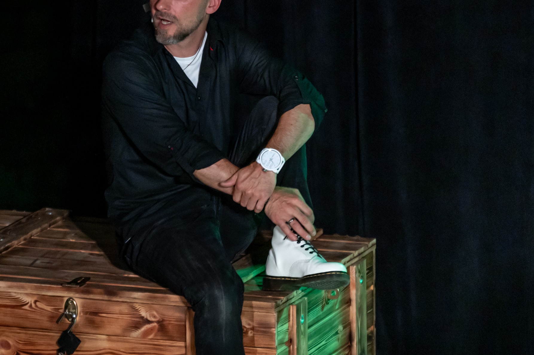 Mężczyzna w czarnej koszuli siedzi na drewnianej skrzyni, opierając stopę w białym bucie na jej krawędzi; widoczny biały zegarek na nadgarstku.