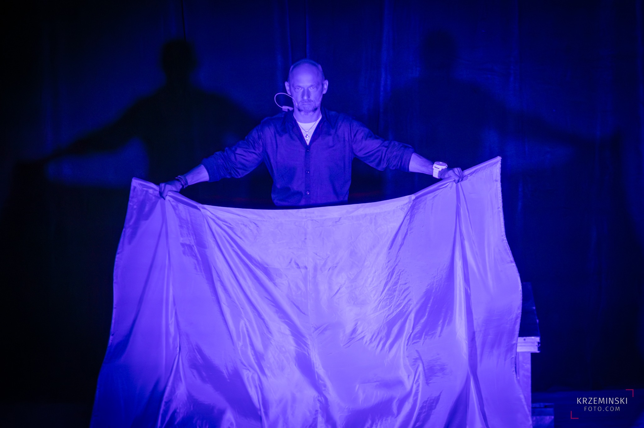 Iluzjonista z Gliwic prezentuje trik z białym płótnem w niebieskim oświetleniu scenicznym, tworząc tajemniczy cień na tle.
