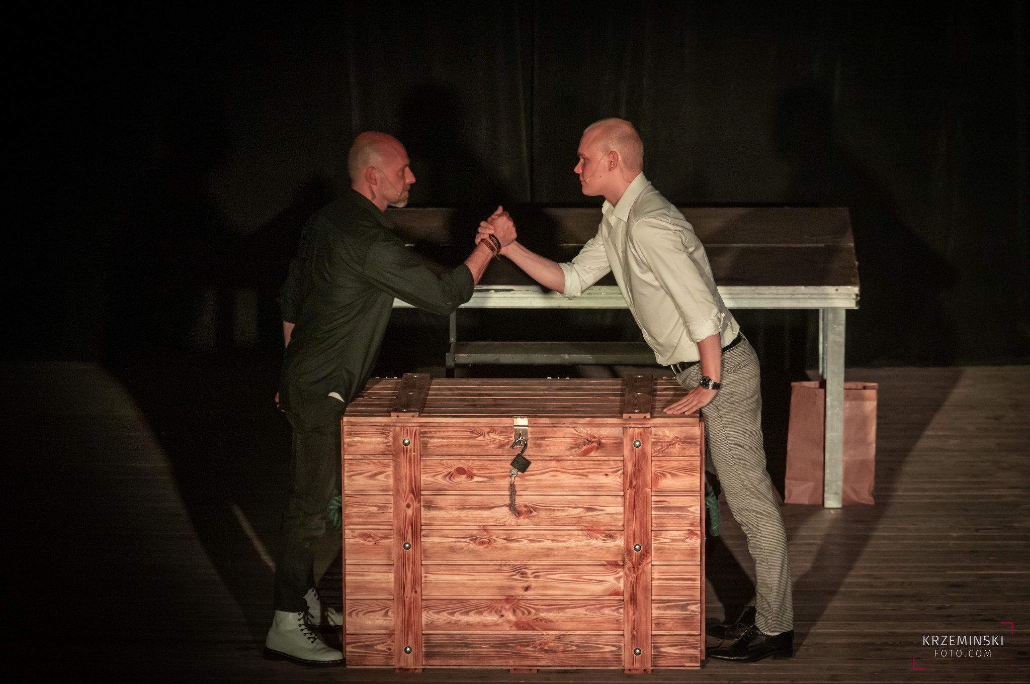 Dwóch mężczyzn w teatralnym oświetleniu, w trakcie scenicznej konfrontacji sił przy drewnianej skrzyni z kluczem.