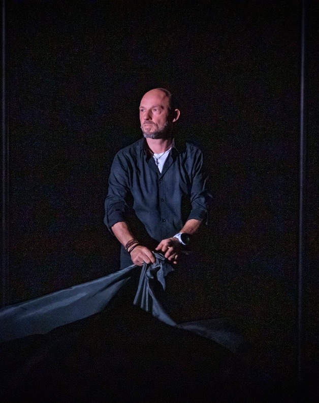 Mężczyzna w czarnej koszuli, z bransoletkami na nadgarstku i zegarkiem, trzymający szary materiał na ciemnym tle, prawdopodobnie podczas występu.