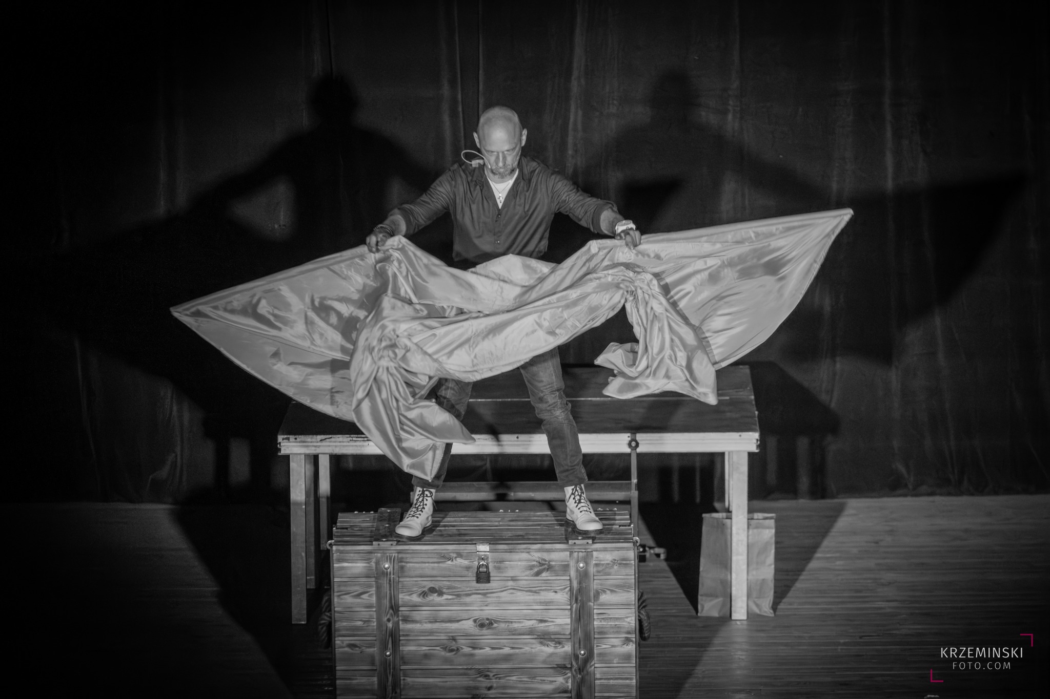 Czarno-białe zdjęcie iluzjonisty rozpościerającego jedwabną tkaninę na scenie, stojącego na drewnianej skrzyni, z widocznym mikroportem i cieniem na tle kurtyny.