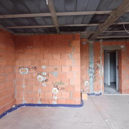 Kruk Michał instalatorstwo elektryczne - Wnętrze w trakcie budowy z widocznymi przewodami elektrycznymi ułożonymi w niebieskich rurkach przy podłodze i wyprowadzonymi punktami na ścianach z czerwonej cegły. Na ścianach widoczne odręczne...