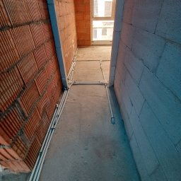 Kruk Michał instalatorstwo elektryczne - Montaż instalacji elektrycznej w surowym wnętrzu: Białe rury prowadzone po podłodze między ścianą z czerwonej cegły dziurawki a ścianą z szarego betonu komórkowego, widok na okno w tle.