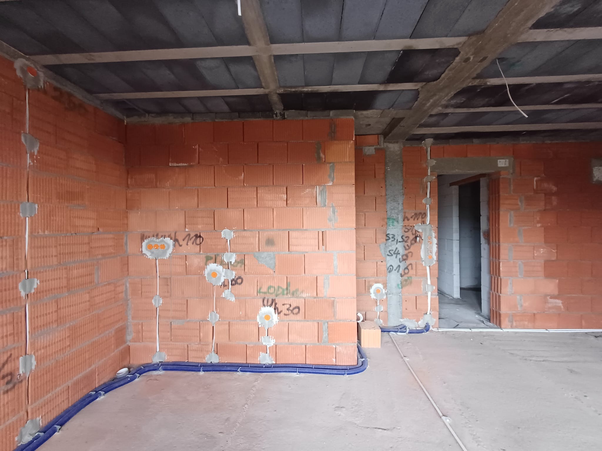 Wnętrze w trakcie budowy z widocznymi przewodami elektrycznymi ułożonymi w niebieskich rurkach przy podłodze i wyprowadzonymi punktami na ścianach z czerwonej cegły. Na ścianach widoczne odręczne...