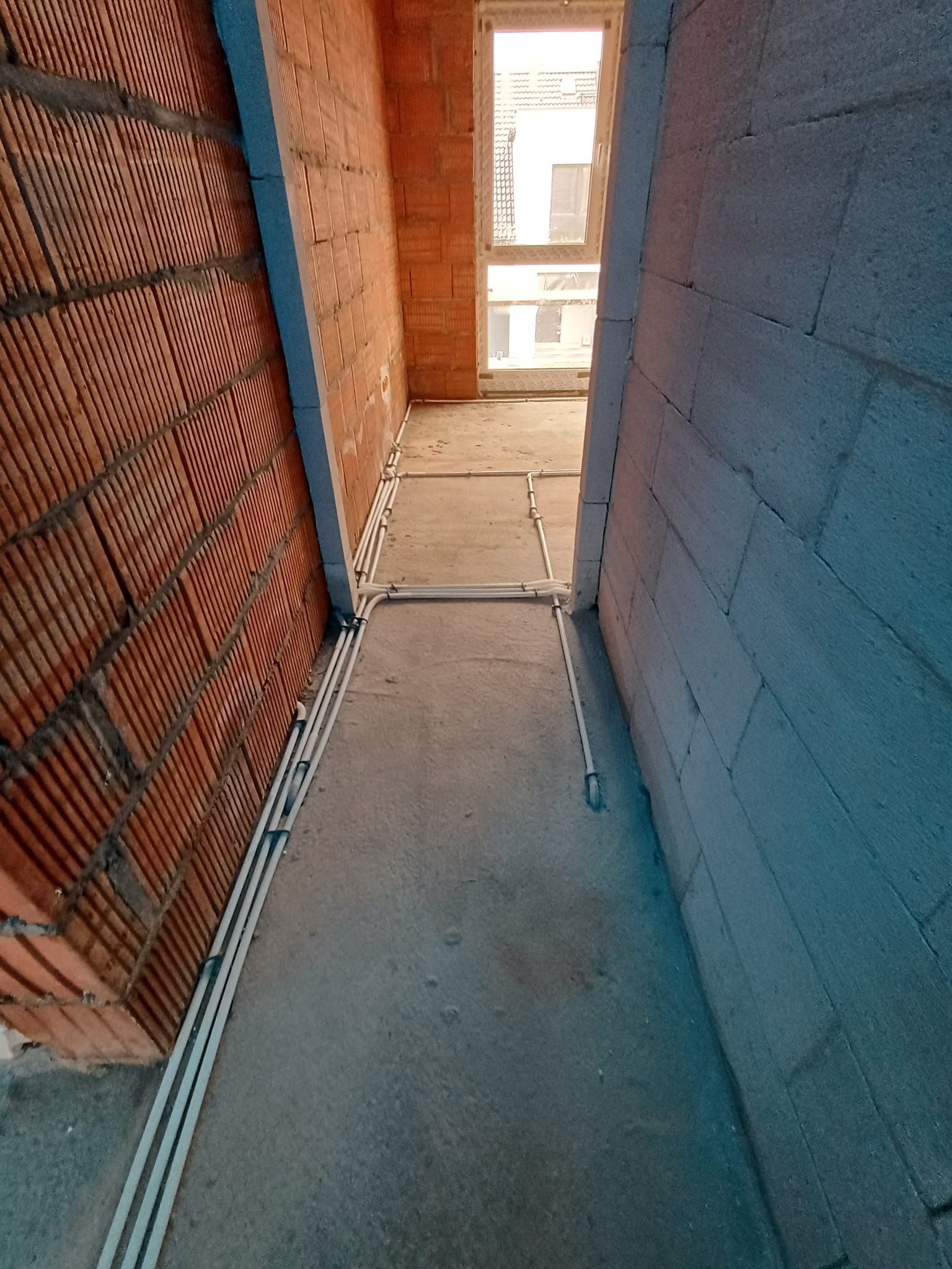 Montaż instalacji elektrycznej w surowym wnętrzu: Białe rury prowadzone po podłodze między ścianą z czerwonej cegły dziurawki a ścianą z szarego betonu komórkowego, widok na okno w tle.