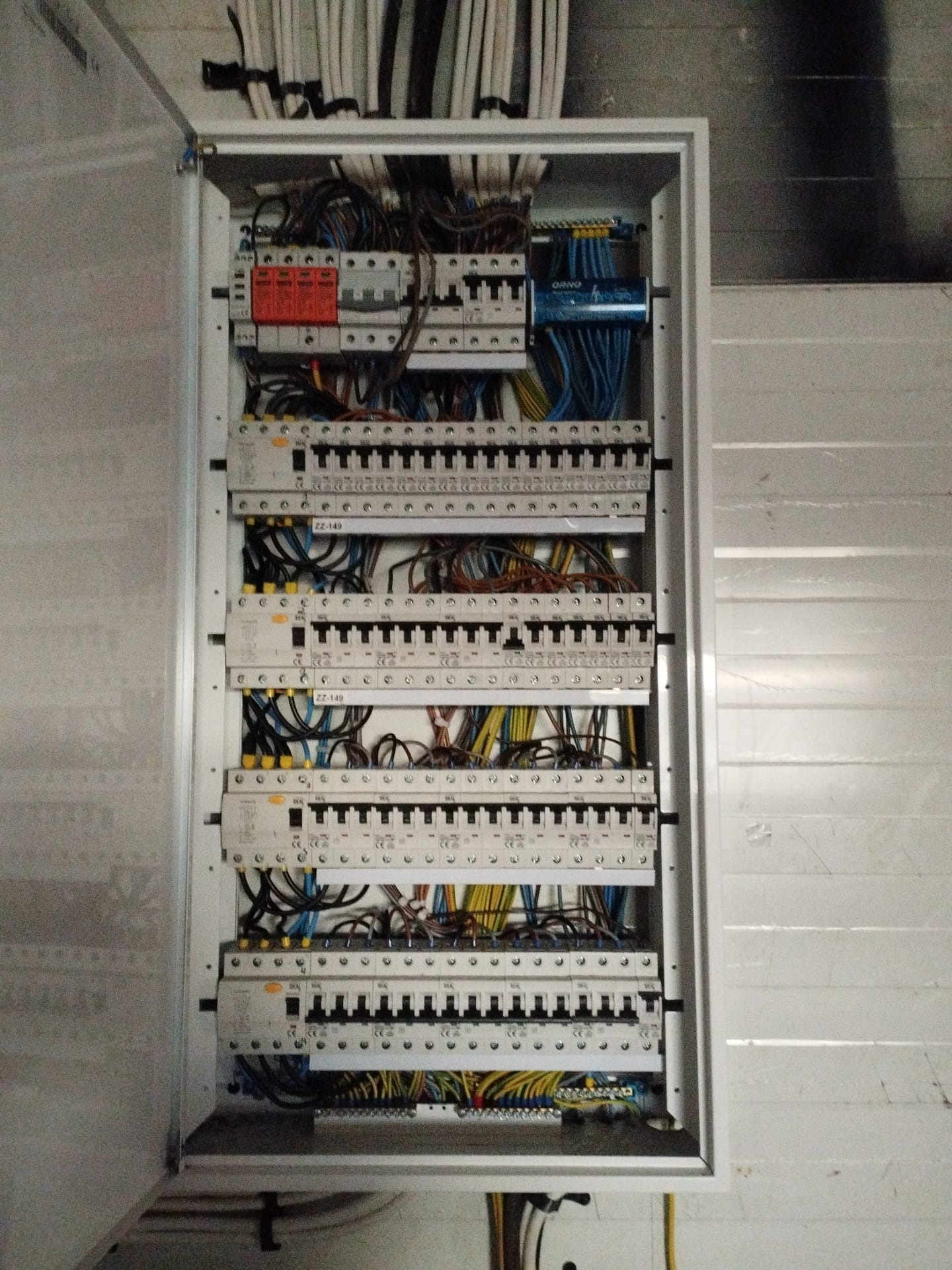 Otwarta skrzynka elektryczna z widocznymi bezpiecznikami, przewodami w różnych kolorach i elementami zabezpieczeń przepięciowych, zamontowana na białej ścianie z poziomymi panelami.