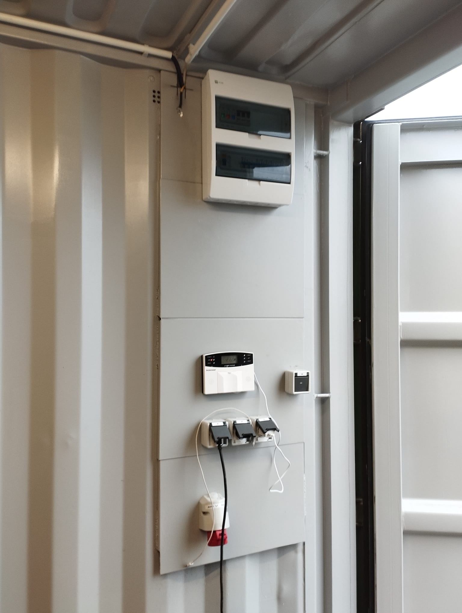 Wnętrze kontenera z zamontowaną białą skrzynką rozdzielczą, panelem sterowania, gniazdkami i przewodem z czerwoną końcówką. Widoczne kable i rurki instalacyjne.