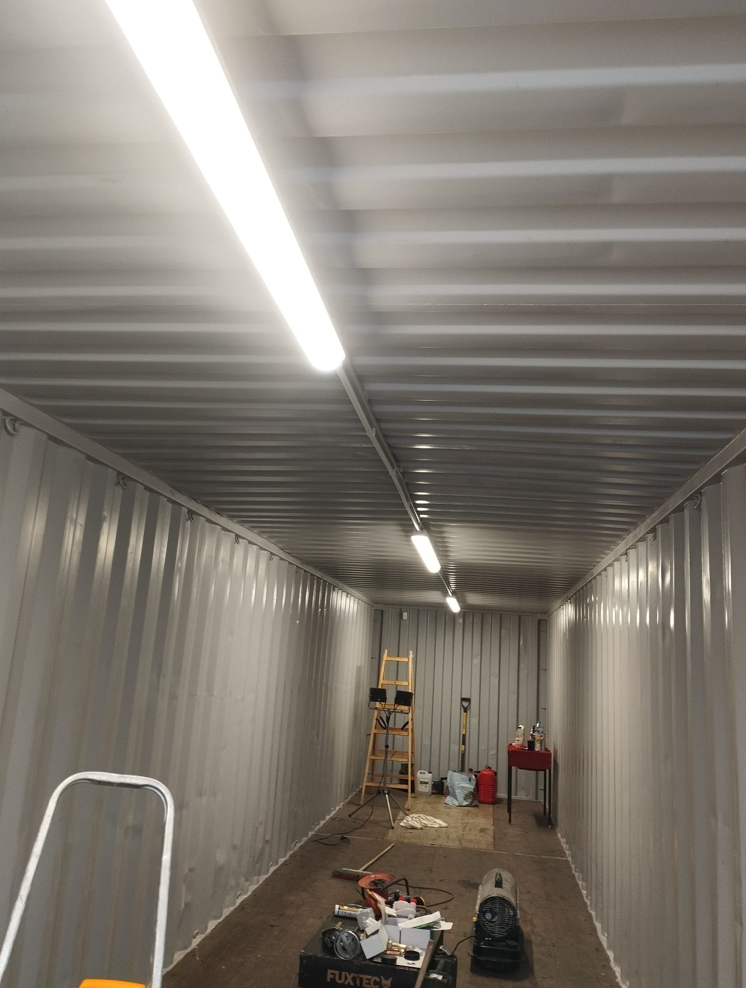 Wnętrze kontenera z zamontowanymi lampami LED na suficie, widoczna drabina, narzędzia i nagrzewnica na podłodze, surowe wykończenie ścian i sufitu.