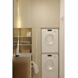 Nowoczesna pralnia z pralką i suszarką w zabudowie, drewniane szafki z półkami, dekoracje, podłoga w odcieniach beżu i szarości.